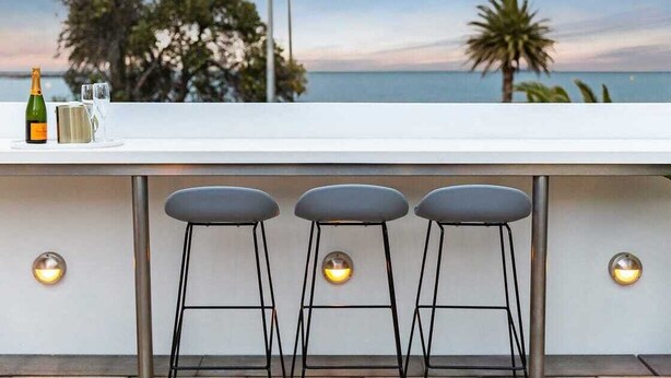Zona do bar com vista para o mar, três banquetas, uma garrafa de champanhe e duas taças.