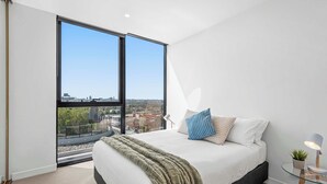 A modern bedroom with a large window, a bed with a blanket, pillows, and a bedside lamp. - Executive apt. +views +parking in beachside resort (Port Melbourne)