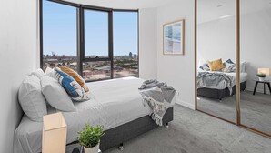 A bedroom with a large window, a bed, a mirror, and a view of the cityscape. - 3BD beachside resort with parking, views and pool (Port Melbourne)