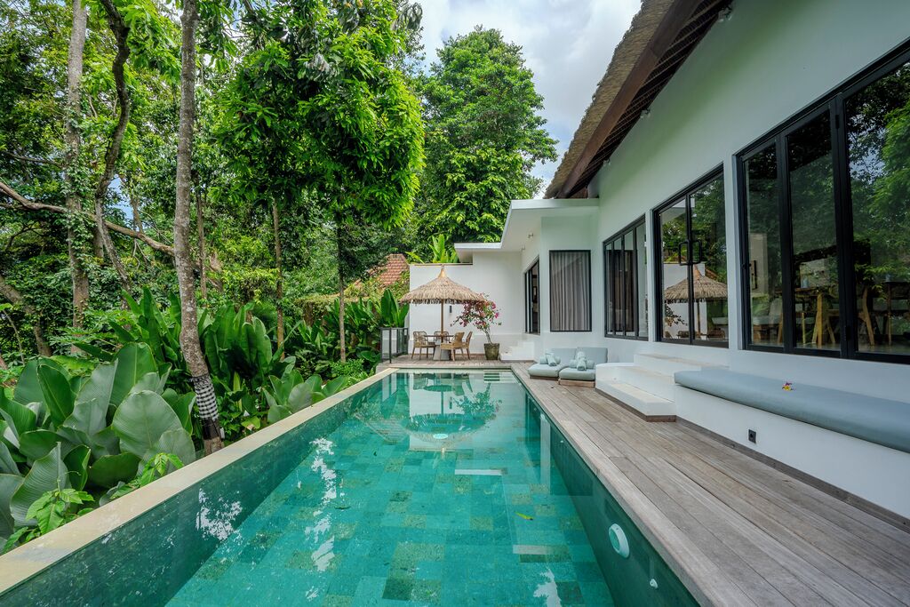 A modern pool area with a wooden deck, surrounded by lush greenery and a white building with large windows.