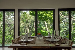 Mesa de comedor lista para cuatro personas con vista a la vegetación exuberante a través de amplios ventanales.