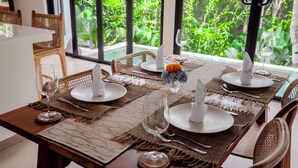 A dining table set with placemats, plates, and cutlery, with a view of lush greenery through large windows. - Infinity Pool Villa with Jungle & River View (Tabanan)