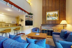 Ein Wohnzimmer mit blauen Sofas, einem Holztisch, einem Kamin und einer Küchenzeile mit weißen Schränken und einer Mikrowelle.