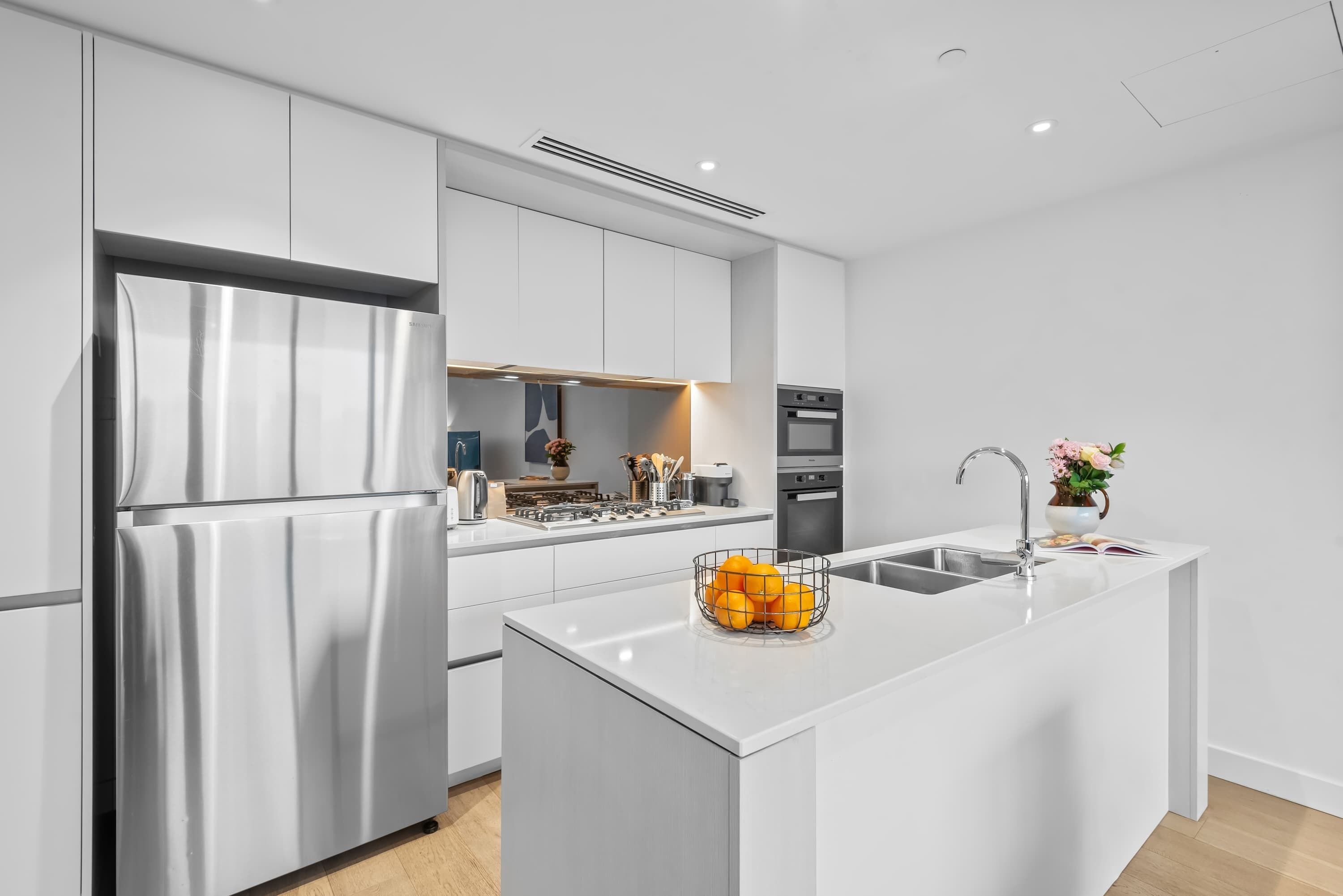 A modern kitchen with white cabinetry, a stainless steel refrigerator, a built-in oven, and a kitchen island with a fruit bowl.