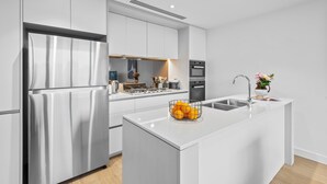 A modern kitchen with white cabinetry, a stainless steel refrigerator, a built-in oven, and a kitchen island with a fruit bowl. - Luxe Docklands Townhouse with 2 Carspaces +Views! (Docklands)