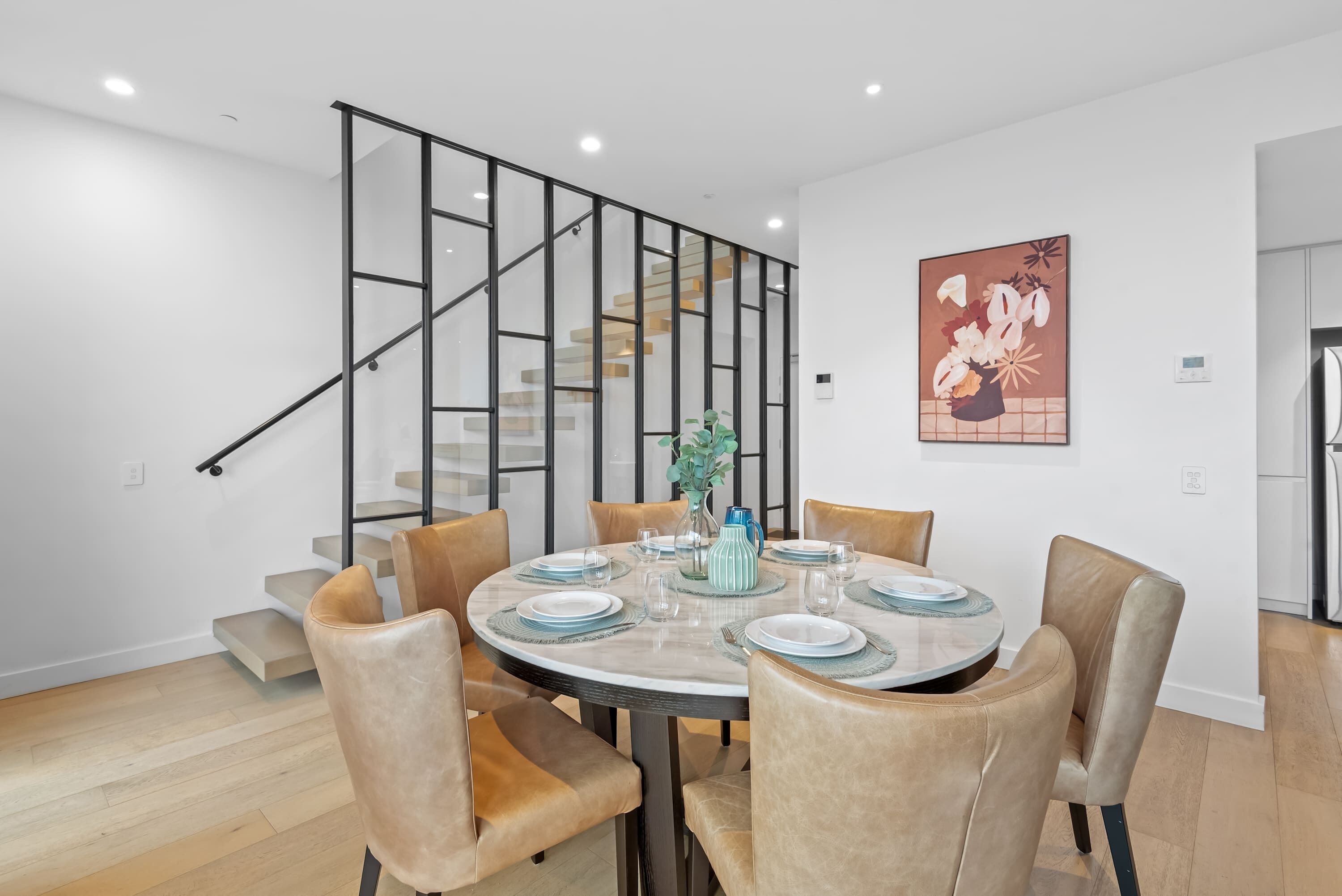 A modern dining area with a round table, six chairs, and a staircase with glass railings.