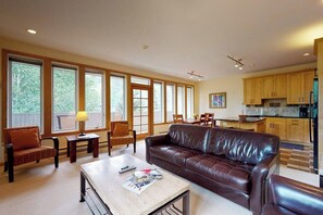 A spacious living room with a leather sofa, a coffee table, and a dining area with a table and chairs. Large windows provide ample natural light. - Bright Mountain Retreat w/Shared Pool & Hot Tub & Mountain Views from Balcony (Vail)