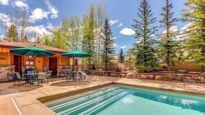 A pool area with a stone deck, outdoor seating, and a wooden building with a green umbrella. - Bright Mountain Retreat w/Shared Pool & Hot Tub & Mountain Views from Balcony (Vail)