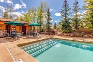 A pool area with a stone deck, outdoor seating, and a wooden building with a green umbrella. - Bright Mountain Retreat w/Shared Pool & Hot Tub & Mountain Views from Balcony (Vail)