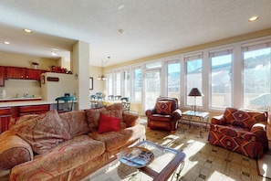 A living room with a sofa, armchair, and coffee table, a kitchen area, and large windows with a view. - Western-inspired condo w/ wood fireplace, full kitchen, & shared hot tub & pool (Vail)