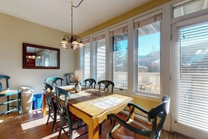 A dining area with a wooden table, chairs, a hanging light fixture, and large windows with blinds. - Western-inspired condo w/ wood fireplace, full kitchen, & shared hot tub & pool (Vail)