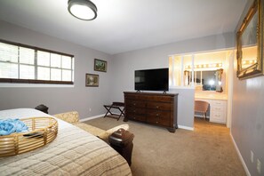 A bedroom with a bed, dresser, television, vanity with mirror, and window with blinds.