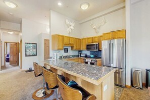A modern kitchen with wooden cabinets, granite countertops, a stainless steel refrigerator, and a microwave. - Mtn-view condo next to lifts with heated outdoor pool, hot tub, sauna, fireplace (Vail)