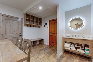 Ein Zimmer mit Esstisch, Stühlen, einer Bank, einer Holztür, einem Wandregal, einem Spiegel mit Beleuchtung sowie einem Badezimmer mit Waschbecken und Handtuchablage.