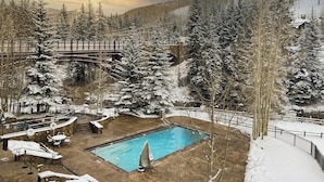 Ein verschneiter Außenbereich mit Schwimmbad, Liegestühlen und einer Holzbrücke.