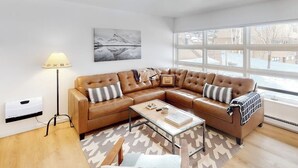 Een moderne woonkamer met een bruine leren hoekbank, een glazen salontafel, een vloerlamp en een groot raam met uitzicht op een besneeerd landschap.