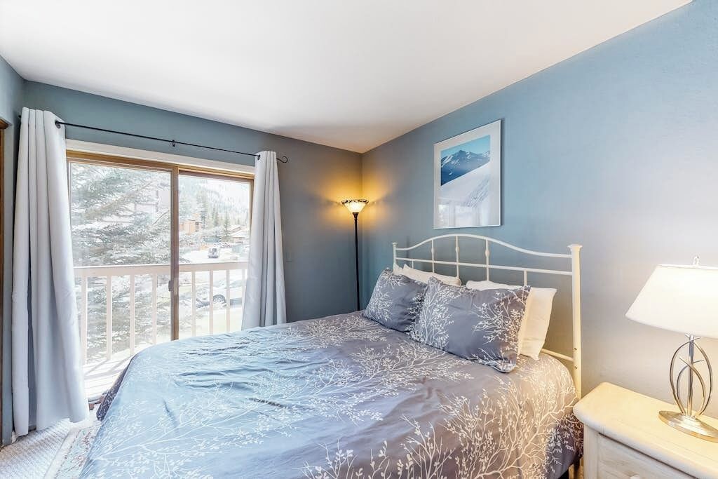 A bedroom with a bed, a nightstand, a lamp, a window with curtains, and a wall-mounted picture.