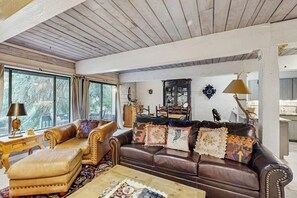 Ein gemütliches Wohnzimmer mit einem braunen Ledersofa, dazu passendem Hocker und einem Couchtisch. Es gibt eine Holzbalkendecke, ein Fenster mit Vorhängen und eine Lampe auf einem Beistelltisch.