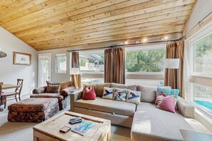 Ein gemütliches Wohnzimmer mit Holzbalkendecke, einer Sofagarnitur, einem Couchtisch und großen Fenstern mit Vorhängen.