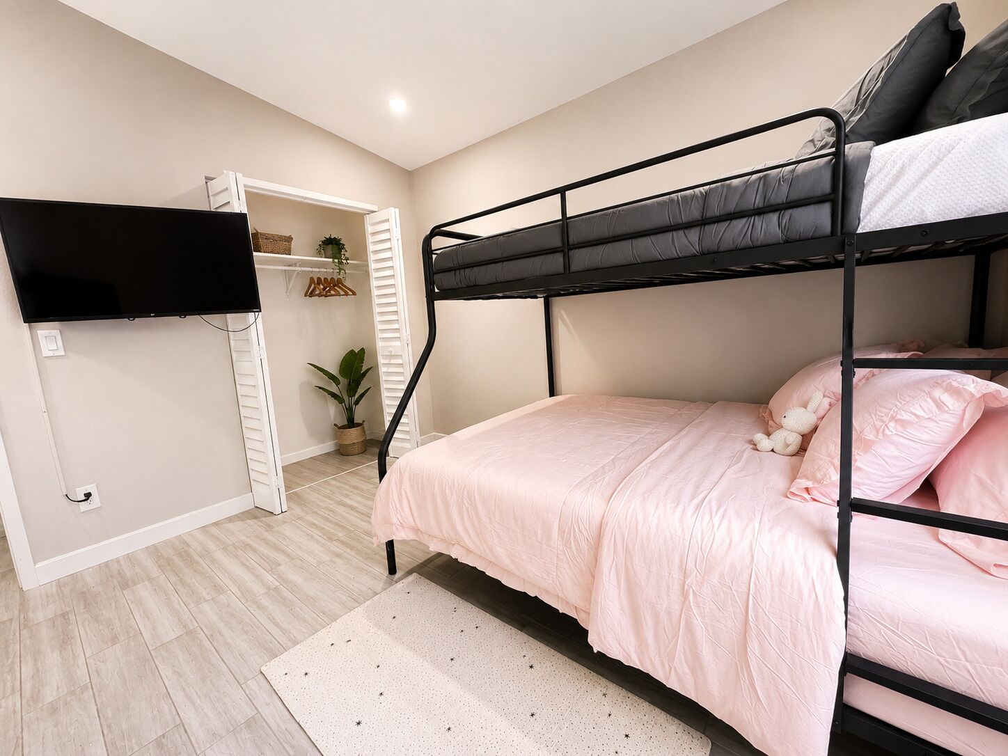 A bunk bed with pink bedding, a television mounted on the wall, a closet with white doors, and a potted plant.