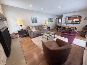 A living room with a sofa, armchair, coffee table, and fireplace. There is a dining area with a table and chairs. The kitchen is visible in the background. - Cozy East Vail condo - Mountain view, Hot Tub, Pool & W/D - Near Ski shuttle (Vail)