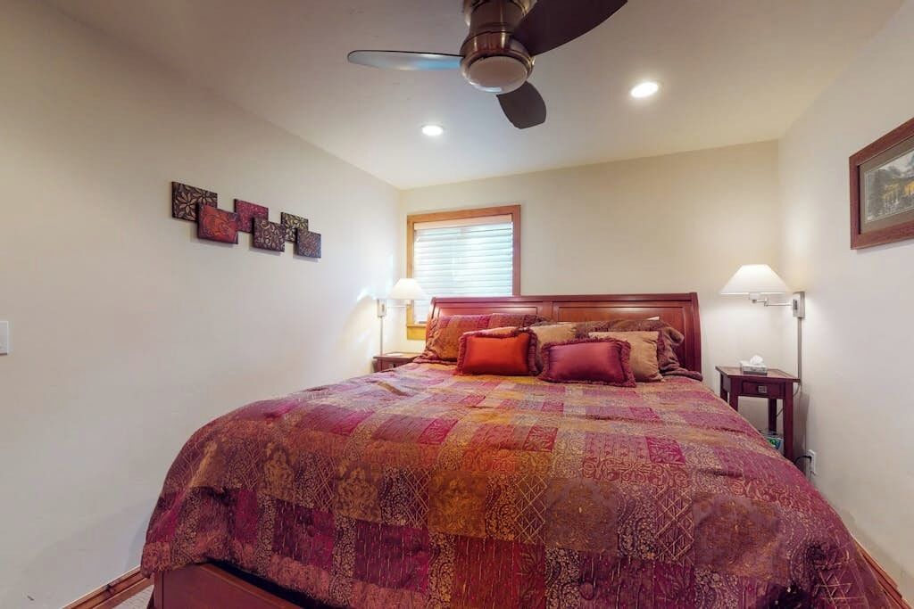 Un dormitorio con cama, ventilador de techo, ventana y lámparas fijadas a la pared.