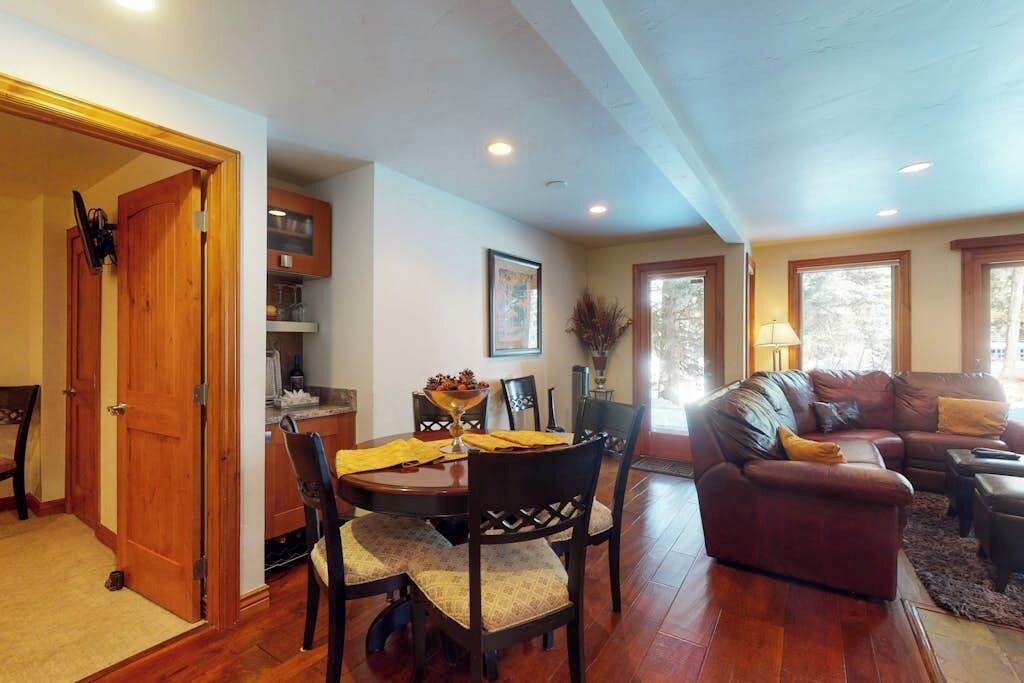 A dining area with a wooden table and chairs, a living room with a leather sofa, and a kitchen with cabinets and a window.