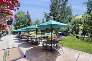 Un espace extérieur aménagé en terrasse, avec des tables et des chaises, des parasols verts et une piscine en arrière-plan.