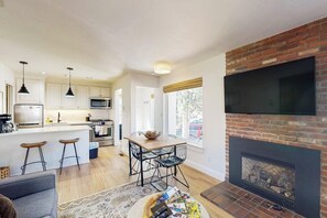Eine moderne Küche mit weißen Schränken, einem Essbereich mit Holztisch und Metallstühlen sowie einem Kamin aus Backstein, über dem sich ein Fernseher befindet.