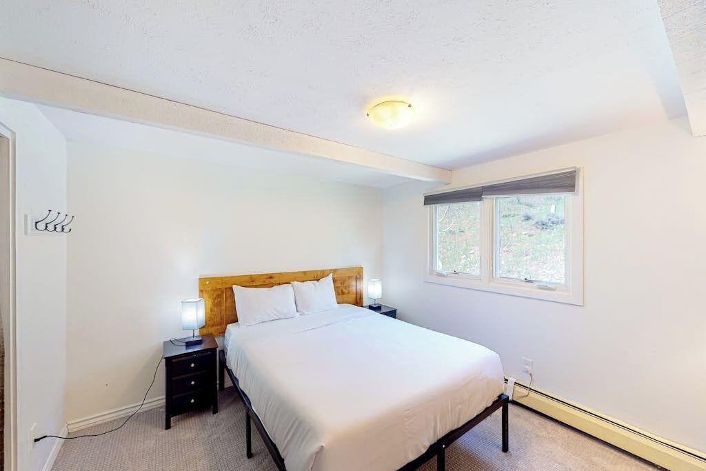 A bedroom with a bed, a nightstand, a lamp, a window, and a ceiling light.
