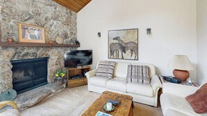 A living room with a stone fireplace, a white sofa, a coffee table, and a television. - Quiet, multi-level townhome with great location & mountain views (Vail)