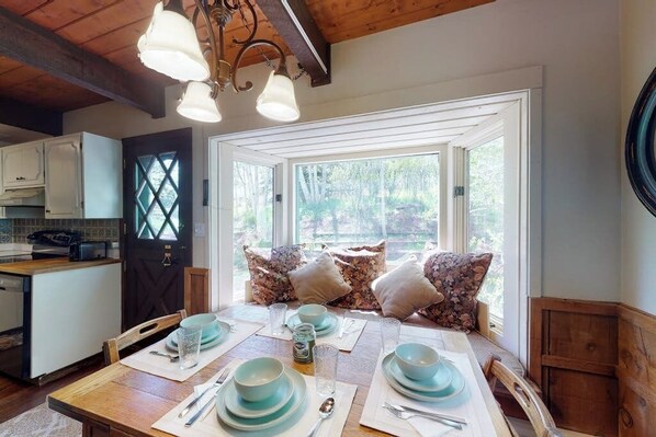 A dining area with a wooden table set for four, a window with a view of trees, and a kitchen in the background. - Quiet mountain-view condo with patio, fireplace & grill - walk to Vail bus (Vail)