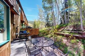 A wooden building with a patio area featuring a round table and chairs, a grill, and a stack of firewood. - Quiet mountain-view condo with patio, fireplace & grill - walk to Vail bus (Vail)