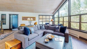 A spacious living room with a large sectional sofa, a coffee table, and a view through large windows. - Mountain-view home with private hot tub, solarium & fireplace - steps to shuttle (Vail)