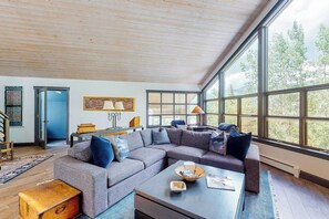 A spacious living room with a large sectional sofa, a coffee table, and a view through large windows. - Mountain-view home with private hot tub, solarium & fireplace - steps to shuttle (Vail)