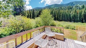 A wooden deck with white metal chairs and a wicker chair, overlooking a forested mountain area. - Mountain-view home with private hot tub, solarium & fireplace - steps to shuttle (Vail)