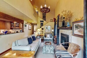 A cozy living room with a sofa, ottoman, and chairs, a fireplace, and a kitchen area with wooden cabinets and a hanging light fixture. - Charming home with private hot tub, fireplace, washer/dryer, and central AC (Vail)
