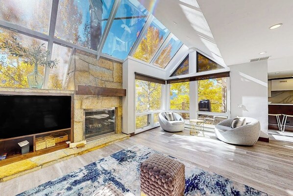 A modern living room with a fireplace, large windows, and a flat-screen TV. - Three-story townhouse with balcony, mountain views, fireplace, grill, & W/D (Vail)