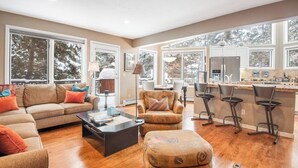 Un salón amplio con sofá, reposapiés y mesa de centro, una cocina con refrigerador y taburetes de bar, y ventanas grandes con vistas a árboles.