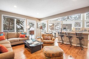 A spacious living room with a sofa, ottoman, and coffee table, a kitchen with a refrigerator and bar stools, and large windows offering a view of trees. - Incredible 3BR mountain view -  steps to ski bus - fireplaces (Vail)