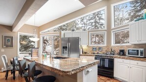 Una cocina con encimera de granito, armarios blancos, un refrigerador de acero inoxidable y un ventanal con vistas a árboles.
