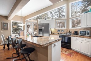 A kitchen with a granite countertop, white cabinets, a stainless steel refrigerator, and a large window overlooking trees. - Incredible 3BR mountain view -  steps to ski bus - fireplaces (Vail)