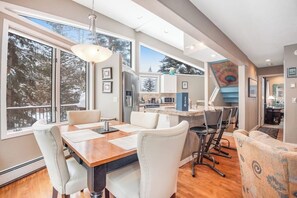 A modern kitchen with a large island, a dining area with a wooden table and chairs, and a view of the outdoors through large windows. - Incredible 3BR mountain view -  steps to ski bus - fireplaces (Vail)