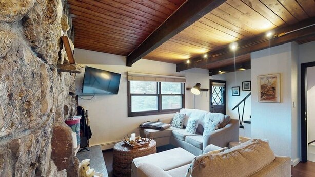 A cozy living room with a stone wall, wooden ceiling beams, a flat-screen TV, a sofa, a coffee table, and a window. - Remodeled, mountain view townhome with patio, fireplace & great location (Vail)