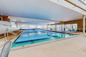 An indoor swimming pool with clear glass walls, a ceiling with linear patterns, and a seating area with tables and chairs. - Rustic condo with resort amenities, sweeping views, and wood-burning fireplace (Vail)