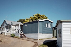 Een rij mobiele huizen met een veranda, een kleine trap en een grote boom op de achtergrond.