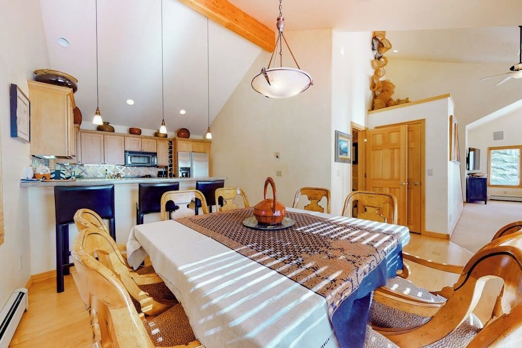 Ein Essbereich mit Tisch für vier Personen, Holzsesseln, einer Hängelampe und einer Küche im Hintergrund.