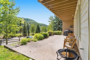 A covered porch with outdoor seating and a grill, overlooking a landscaped area with trees and a mountain. - Sunlit, mountain view condo with a fireplace plus on-site pool & hot tub (Vail)