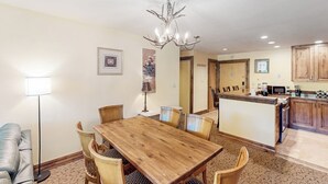 A dining area with a wooden table and chairs, a chandelier, and a kitchen in the background. - Charming condo with resort amenities, sweeping views, & wood-burning fireplace (Vail)