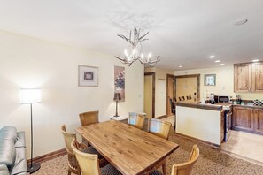 A dining area with a wooden table and chairs, a chandelier, and a kitchen in the background. - Charming condo with resort amenities, sweeping views, & wood-burning fireplace (Vail)